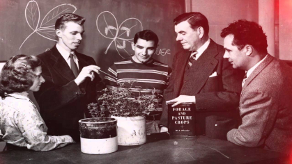 A vintage photo of professors and students inspecting a potted plant in a classroom.